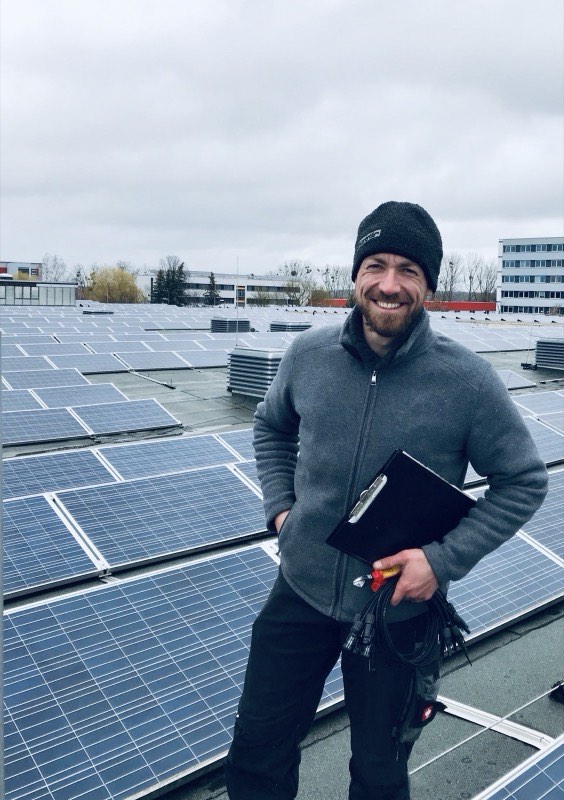 Roman Schladebusch mit Mütze und Klemmbrett auf einem Flachdach voller Solarmodule